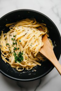 Top-down view of fettuccine pasta being tossed into the prepared creamy mushroom sauce inside a black skillet, placed on a white marble countertop. The pasta is well coated in the sauce and topped with freshly chopped parsley for a fresh finish. This final step brings all the components together into one delicious dish.