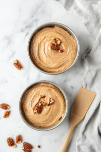 Two round cake pans are filled with smooth, evenly spread carrot cake batter, sitting over a clean white marble countertop. The batter shows bits of grated carrots and pecans, and the tops have been leveled with a rubber spatula. This step ensures uniform baking and prevents the cake from rising unevenly in the oven.