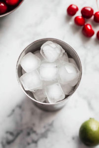A top-down view of the second step in the Mistletoe Martini recipe, showing clear ice cubes being added to a silver cocktail shaker already filled with vodka, cherry brandy, lime juice, and simple syrup. The shaker sits on a white marble countertop, surrounded by a jigger and small empty ingredient bowls. This step visually emphasizes the importance of chilling the cocktail mixture before serving. The clean and organized setup reflects a professional and home-friendly approach to cocktail making, perfect for visual storytelling in recipes.