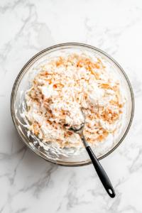 After a few hours in the refrigerator, the coleslaw is being stirred one final time in a large glass bowl on a white marble surface. This step ensures that the creamy dressing is evenly distributed and the texture is consistent throughout. The cabbage and carrots look vibrant and coated in the rich dressing.
