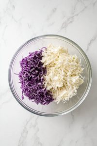 Finely shredded white and red cabbage are placed into a large transparent glass mixing bowl sitting on a white marble kitchen countertop. The sharp knife and wooden cutting board used for chopping are beside it. The fresh shreds of cabbage create a vibrant contrast, with the white cabbage looking crisp and the purple cabbage adding rich color.