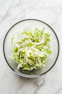 A top-down view of a green cabbage being finely sliced on a wooden cutting board using a sharp stainless steel knife. The sliced cabbage is being added into a large clear glass mixing bowl placed on a white marble countertop. The setup is neat, clean, and highlights the thin, crisp texture of the cabbage ready for salad preparation.