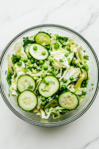 Top-down view of the complete cabbage cucumber sweet pea salad served in a large clear glass bowl on a white marble countertop. The vibrant green cabbage, crisp cucumber slices, sweet peas, and chopped green onions are evenly coated in a light vinegar and olive oil dressing, creating a fresh and colorful side dish perfect for warm days.