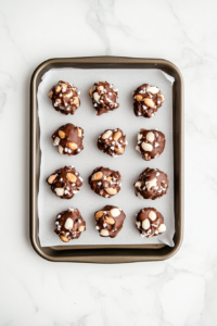 Top-down view of finished chocolate peanut clusters arranged neatly on a sheet of parchment paper placed over a baking tray. The glossy chocolate covers each peanut-filled cluster, giving them a rich and indulgent appearance. These homemade treats are cooling and ready to enjoy, showcasing the perfect blend of white and semisweet chocolate with roasted peanuts.