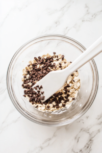 Mini dairy-free chocolate chips are being gently folded into the sticky oat and cashew butter mixture with a white silicone spatula. The chips are clearly visible against the textured dough. The clean white marble background and minimal setup allow the viewer to focus on the incorporation of the final sweet ingredient that gives these bites their delicious pop of flavor.