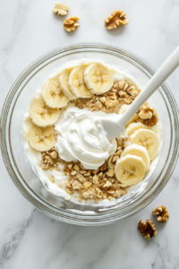 Top-down view of sour cream, sliced bananas, and chopped walnuts being gently folded into the banana bread batter inside a clear glass bowl using a white spatula. The mixing bowl rests on a white marble surface. The creamy texture of the batter is visible with banana slices and walnut chunks partially blended in, emphasizing the flavor-packed and moist nature of the recipe.