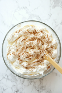 Top-down view of whipped cream being gently folded into the chocolate cream cheese mixture inside a clear glass bowl. A rubber spatula blends the airy whipped cream with the rich chocolate filling. This step ensures a fluffy, mousse-like texture. The scene is set on a white marble countertop with minimal, clean background elements.