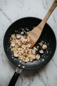 Freshly sliced button mushrooms are being sautéed in the butter and garlic mixture in a black skillet placed on a white marble countertop. The mushrooms have started to soften and shrink, absorbing the flavorful base. The cooking process is simple yet vibrant, showcasing the rich texture and early transformation of the mushrooms.