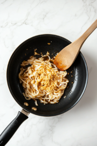 A top-down view of finely ground ginger sprinkled onto glossy teriyaki noodles in a black nonstick pan. A wooden spoon is stirring everything together to blend the flavors. The ginger adds a warm, slightly spicy kick to the dish, enhancing the depth of the teriyaki sauce and balancing sweetness.