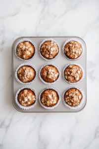 Top-down view of golden brown banana oat muffins with melted chocolate chips, arranged in white paper liners in a muffin pan. The muffins look soft and fluffy with a slightly crisp top. They are placed on a clean white marble countertop, and the scene is well-lit with a warm, homemade feel.