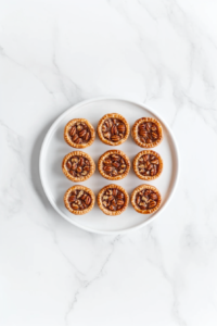 A top-down view of freshly baked golden mini pecan pies arranged on a white ceramic plate over a clean white marble countertop. The pies have a shiny, caramelized pecan topping and a flaky crust, capturing the homemade warmth and richness of this classic dessert. Perfectly baked and ready to enjoy with family or guests.