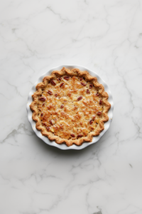 Top-down view of a freshly baked homemade apple pecan pie sitting in a white ceramic 9-inch pie dish on a white marble countertop. The pie has a golden flaky crust and is topped with a rich layer of glistening chopped pecans caramelized with brown sugar and margarine. The scene is clean, rustic, and inviting.