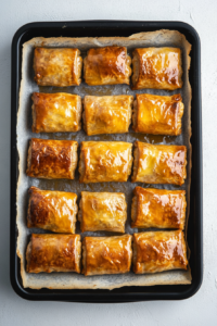 Golden brown sausage rolls rest on a black baking tray lined with parchment paper, fresh out of the oven. The pastry is crisp, flaky, and evenly baked. A small dish of ketchup sits beside the tray, ready for dipping. The rolls are slightly cooling before serving, ensuring the best texture and taste. The clean background keeps the focus on the perfectly baked sausage rolls.