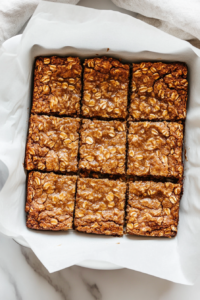 Top-down image showing perfectly baked gingerbread oat bars sliced inside a parchment-lined 8-inch baking tin on a white marble countertop. The bars are golden brown with a shiny top and soft texture. Bits of candied ginger are visible, enhancing the homemade appearance. The clean setup highlights the rustic charm of this spiced dessert.