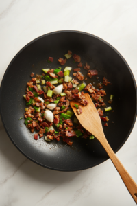 A black frying pan sits on a white marble countertop, filled with sizzling minced bacon, finely chopped brown onion, celery, and minced garlic. The ingredients are cooking in olive oil, turning golden and fragrant. A wooden spatula rests on the edge of the pan, showing the process of sautéing until softened. The mixture will add a rich, savory taste to the sausage rolls.