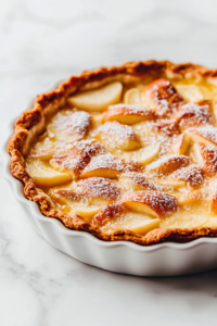 A fully baked apple custard tart beautifully presented in a white tart pan with removable bottom. The tart features golden-brown edges, perfectly layered apple slices, and a glossy glaze from apple jelly. Lightly dusted with confectioners’ sugar, the tart is placed on a white marble countertop, ready to serve. This image highlights the final baked product with a rustic yet elegant appearance, showcasing the results of the recipe in a clean and appetizing setup.