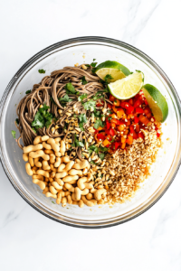 This image captures the final garnish step, with coarsely chopped salted peanuts sprinkled generously over the top of the soba noodle salad. A fresh lime wedge sits to the side, ready to be squeezed over. The noodles, carrots, and cilantro are well-tossed and coated in the dressing, making the dish look both nutritious and inviting, ready for serving.