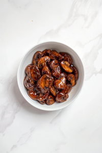 A top-down view of glossy garlic butter mushrooms served in a clean white bowl, placed on a white marble countertop. The mushrooms are perfectly cooked and coated in a rich brown oyster sauce glaze. The dish looks warm, flavorful, and comforting, ideal to pair with Instant Pot Calrose Rice or crispy Breaded Chinese Chicken.