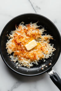 Top-down image of golden hashbrowns cooking in a black nonstick skillet with butter. The hashbrowns have crispy, lightly browned edges and are being sautéed evenly. The skillet is placed on a white marble countertop. This step adds a rich, crispy texture to the breakfast casserole, making it extra hearty and comforting.
