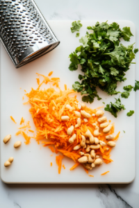 A clean white cutting board on a white marble surface holds three colorful and essential toppings: freshly grated orange carrots, finely chopped green cilantro, and coarsely chopped salted peanuts. A silver grater and knife rest nearby, indicating that the prep is freshly done. These ingredients add crunch, freshness, and a burst of flavor to the soba noodle salad.