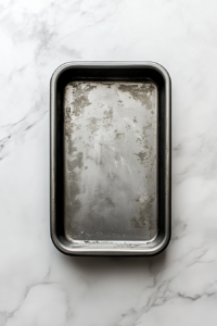 Top-down view of a 9x5 inch metal loaf pan being greased with melted butter using a pastry brush, with an oven thermometer nearby showing 350°F. Everything is placed over a clean white marble countertop. The simple, neat setup includes a small bowl of butter, emphasizing the preparation process before baking banana bread, ensuring proper non-stick results.