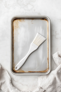 A top-down view of a silver nine by thirteen inch baking pan being greased with a white silicone brush on a white marble countertop. A small bowl of melted butter and a folded white kitchen towel sit nearby. The scene is clean and simple, showing the essential first step to prepare the pan before baking.