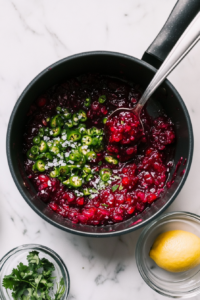 This top-down image shows a vibrant cranberry mixture simmering in a black saucepan as freshly diced green jalapeños, lemon juice, dried cilantro, and salt are added. Each ingredient is visibly blended in, enhancing the flavor complexity. This step adds heat, freshness, herbal notes, and a hint of saltiness to the sweet berry mixture.