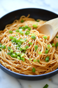 A top-down image of freshly chopped green onions and white sesame seeds being added as garnishes over the hot teriyaki noodles in a black nonstick pan. These optional toppings add texture, freshness, and visual appeal, while also elevating the overall taste with a slight crunch and brightness.