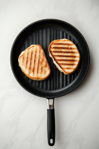 Two assembled four-cheese sandwiches are being grilled in a black non-stick skillet placed on a white marble countertop. One sandwich is being flipped with a black-handled spatula, while the other continues to toast to a golden brown. Melting cheddar and provolone cheese are visible between the crisp slices of artisan bread. This image shows the cooking step in action, capturing the perfect balance between texture and color, helping users visually confirm correct grilling.