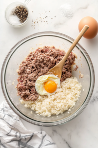 Top-down shot of a glass mixing bowl filled with raw ground beef, chopped onions, partially cooked rice, a cracked egg, salt, and pepper on a white marble countertop. This image highlights the preparation of the flavorful filling used to stuff the cabbage leaves. Everything is evenly mixed, showcasing a rich and well-seasoned base.