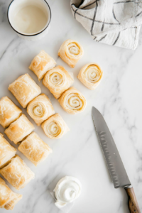 Hands carefully roll the puff pastry over the seasoned pork filling, sealing the edges with whisked egg using a pastry brush. The pastry looks soft and smooth, ensuring a perfect seal. A sharp knife is placed nearby, ready to slice the rolls into smaller pieces. The clean, white marble countertop keeps the focus on this crucial step of shaping the sausage rolls.
