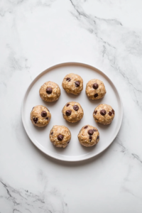 A top-down view of finished no-bake energy bites made with creamy cashew butter, oats, flaxseed, and mini chocolate chips. The bites are perfectly round and arranged on a white ceramic plate, highlighting their chewy texture and speckled chocolate chips. The clean, white marble background enhances the wholesome and homemade look of this protein-rich snack.