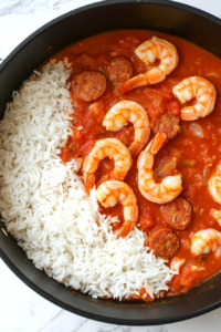 Freshly cooked long-grain white rice is being stirred into the shrimp, sausage, and tomato mixture inside a black pot. A wooden spoon mixes the ingredients, ensuring the fluffy rice absorbs the vibrant red sauce. Shrimp and sausage pieces are evenly distributed, making the dish look flavorful and inviting.