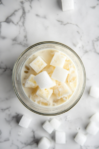 Top-down shot of 8 ice cubes being added to a glass blender already containing a mix of coconut milk, pineapple juice, rum, and sugar. The blender is set on a white marble countertop, with scattered ingredients nearby. The ice adds the chill and texture needed to turn this mix into a smooth, frozen delight.