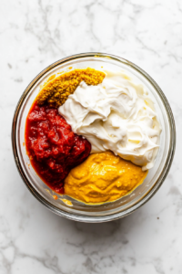 A top-down view of a clear glass mixing bowl containing measured ingredients: mayonnaise, chili sauce, Creole mustard, olive oil, and fresh lemon juice. The bowl is placed on a clean white marble countertop. Each ingredient is clearly distinguishable and freshly added, showcasing the start of a flavorful remoulade sauce preparation using common pantry staples and tangy elements.