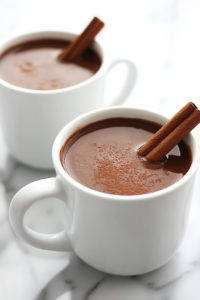 A ladle filled with thick, warm hot chocolate is shown pouring into white heatproof mugs placed on a white marble surface. The silver saucepan is off the heat, and the chocolate inside has a silky finish. This moment captures the final step before serving, emphasizing the cozy and heartwarming appeal of this homemade spiced drink.