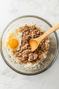 A large glass mixing bowl on a white marble countertop contains raw ground pork, panko breadcrumbs, a cracked egg, fennel seeds, salt, black pepper, and sautéed garlic, onion, celery, and bacon. A wooden spoon is partially submerged in the mixture, combining the ingredients. The background is clean, keeping the focus on the flavorful filling for the sausage rolls.