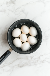 Six large white eggs are submerged in water inside a black saucepan placed on a white marble countertop. The water is beginning to heat as the eggs prepare to boil. This step is crucial for ensuring perfectly cooked eggs with firm whites and creamy yolks, the foundation of a rich and flavorful egg salad.