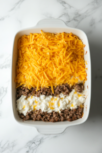 Top-down view of a white baking dish with visible layers of golden hashbrowns, sausage with diced tomatoes and green chiles, and a generous topping of shredded Colby Jack cheese. The dish is halfway filled and sits on a clean white marble countertop. Each ingredient is evenly spread to build flavor and texture.