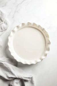 A 9-inch white ceramic pie dish is being carefully lined with a pastry crust on a clean white marble countertop. The edges of the dough hang slightly over the rim, ready to cradle the flavorful apple and pecan filling. This step is essential for creating a firm, buttery base for the pie.