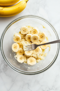 Top-down view of three ripe bananas mashed into a smooth mixture inside a clear glass mixing bowl, placed on a white marble countertop. A silver fork lies nearby. This step is essential for achieving the moist, sweet base of the banana bread, and the texture of the mashed fruit is clearly visible.