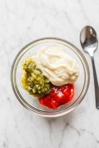 A top-down shot of a glass mixing bowl containing mayonnaise, ketchup, and relish with a fresh addition of Dijon mustard. The mustard, with its yellow hue, sits on top, about to be mixed in. This key step enhances the tangy flavor profile of the burger sauce and adds depth to its creamy base.
