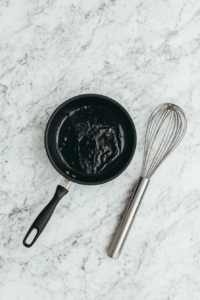 This photo shows a black saucepan on a white marble surface filled with a glossy mixture of melted black treacle, golden syrup, brown sugar, butter, and lard. The ingredients are gently melted and not boiled, creating a rich dark syrup. The warm blend adds depth and sweetness, forming the flavor base of the recipe.