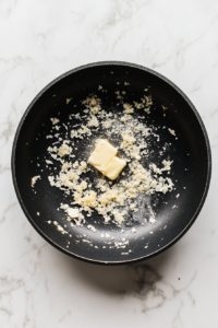 A top-down image of golden sautéed mushrooms in a black skillet with melting unsalted butter and freshly grated garlic added in. The skillet is on a white marble countertop, and the garlic is beginning to release its rich aroma. This step infuses the mushrooms with a fragrant buttery flavor foundation.