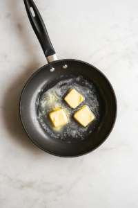 Butter is melting inside a black skillet placed on a bright white marble countertop. The butter is partially melted and slightly foamy, preparing the base for sautéing garlic. The clean and minimal kitchen background makes the process of starting this mushroom dish look simple and easy to follow for home cooks.