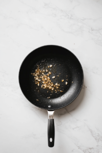 Top-down view of a black skillet placed on a white marble countertop with butter melted inside. Minced garlic is being sautéed and is just starting to turn golden, releasing a rich, aromatic fragrance. A garlic press and wooden spoon are nearby, and the butter is bubbling gently. This step infuses the butter with savory garlic flavor, which forms the base of the delicious pumpkin sauce in this pasta recipe.