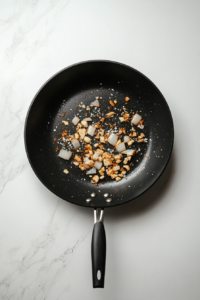 In a black skillet, chopped onions, minced garlic, and fresh ginger are being sautéed in melted butter and sesame oil over a white marble cooktop. The ingredients are lightly browned and fragrant, forming a savory base. This is a key step that infuses the noodles with layers of aromatic flavor and warmth before combining with the sauce.