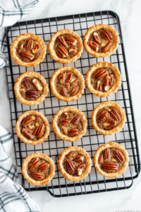 Freshly baked mini pecan pies are placed on a wire rack to cool for 15–20 minutes over a white marble countertop. Cooling allows the filling to fully set and gives the pies a firmer texture for easier handling. The rich aroma of toasted pecans and buttery crust fills the air while they rest.