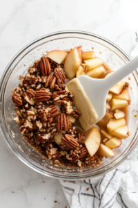 Chopped pecans, packed brown sugar, and melted margarine are being stirred together in a clear glass mixing bowl placed on a white marble countertop. This sweet and nutty mixture will be spread over the apple filling to create a golden, crunchy topping that perfectly complements the tender apple center of the pie.