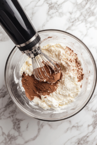 Cream cheese is beaten smooth in a clear glass bowl, with sifted powdered sugar and cocoa powder added for a chocolatey, creamy cheesecake base. This bowl sits on a clean white marble surface, and the electric hand mixer rests nearby. The mixture is rich, velvety, and essential for the dessert’s smooth texture.